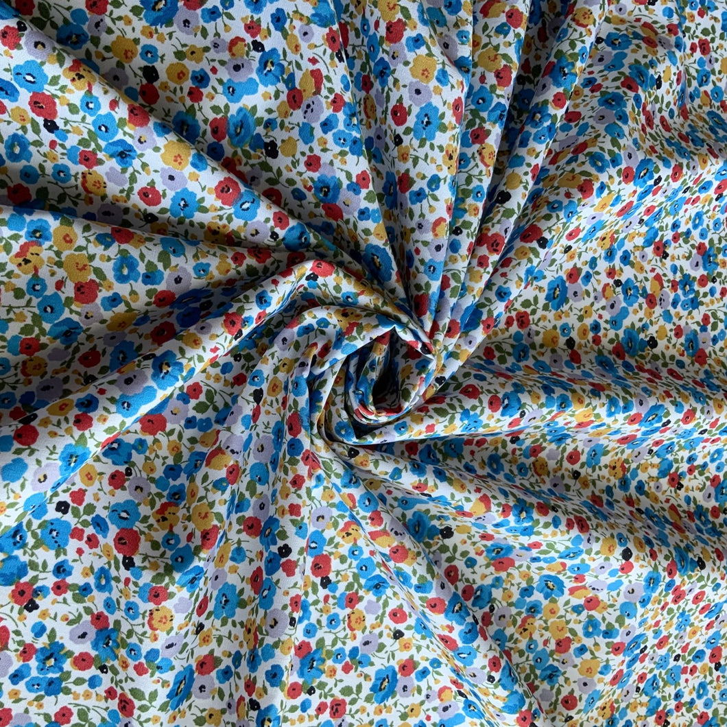 A cotton poplin fabric with a blue themed floral meadow print. The design is a subtle water colour poppy style flowers with colours in yellow, lilac, red and blue against a pale white background. With a crisp, smooth handle and a slight drape, it would be a perfect sewing fabric for dressmaking projects or craft.